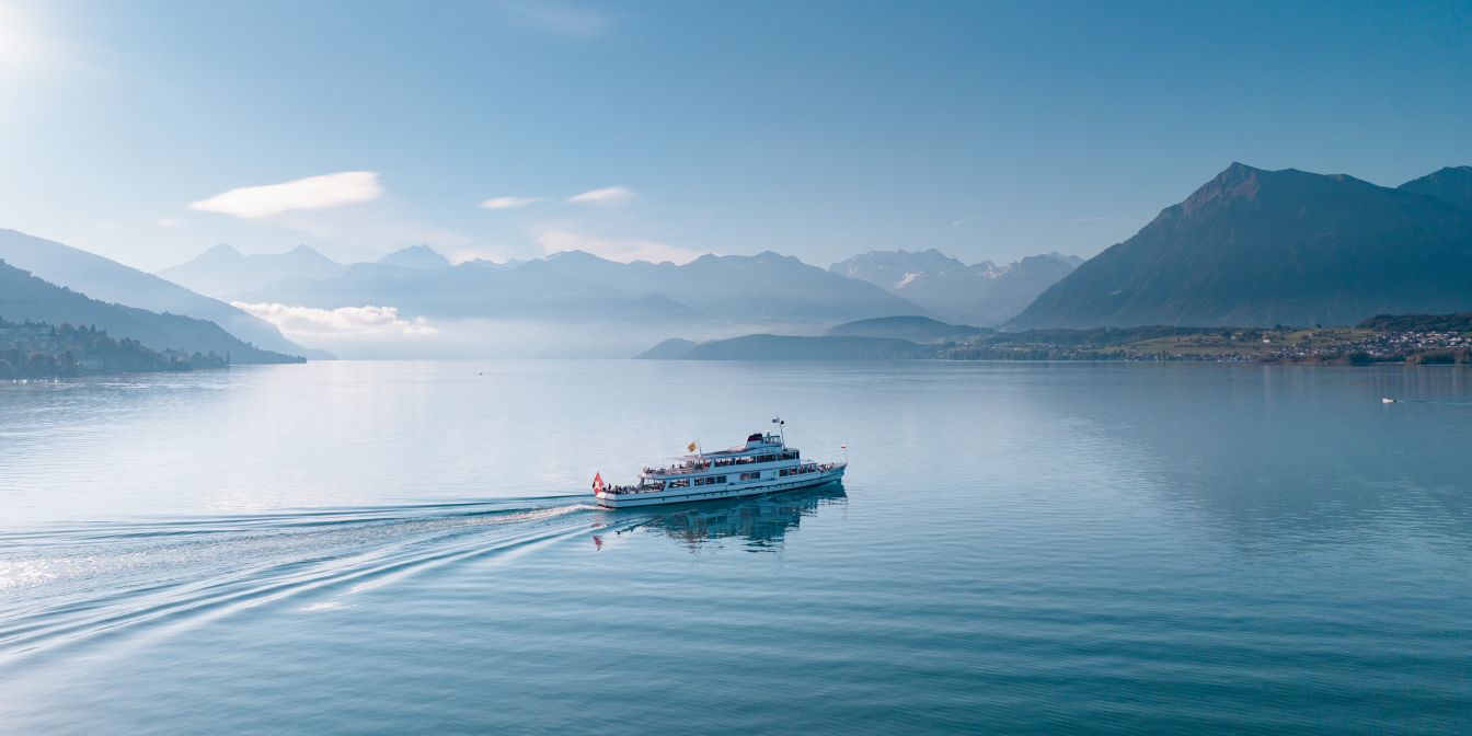 Ein Schiff zieht ruhige Wellen über den Thunersee; im Hintergrund liegen Berge und Nebel – inspirierende Kulisse für Business-Events in der Region. ©BLS Schifffahrt AG Passagierschiff auf dem Thunersee vor Alpenpanorama am Morgen