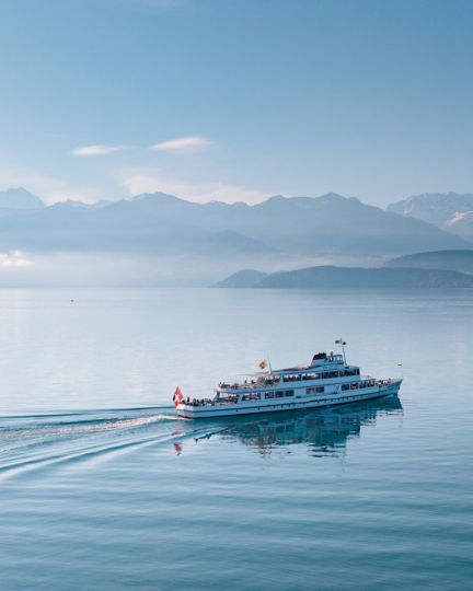 Ein Ausflugsschiff gleitet gemächlich über den ruhigen Thunersee, während sich im Hintergrund majestätische Alpen erheben. Die sanfte Morgensonne sorgt für eine beruhigende Atmosphäre und lässt das Wasser und die umliegende Landschaft in zarten Blautönen leuchten. ©BLS Schifffahrt AG. Schifffahrt auf dem Thunersee
