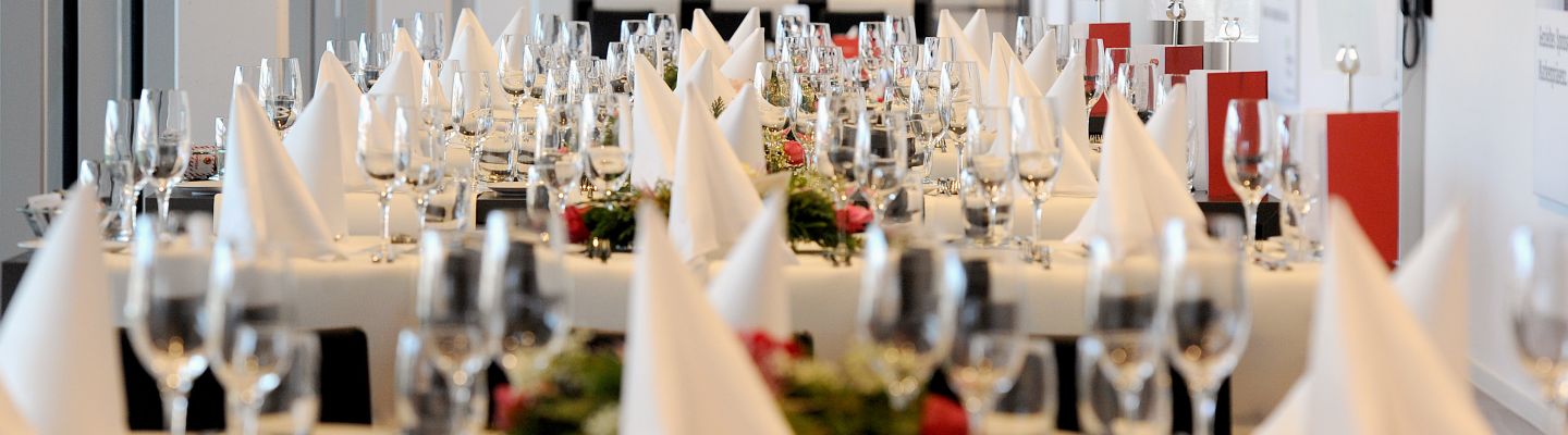 Lange Tischreihen für ein Business-Dinner, mit weissen Servietten, Gläsern und Blumendeko – stilvolles Event-Setting im Tagungsraum. ©Stockhornarena Festlich gedeckte Banketttische mit Serviettenfaltungen und Weingläsern
