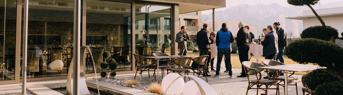 Eine Gruppe von Geschäftsleuten trifft sich auf einer stilvollen Terrasse mit gepflegten Sitzbereichen und Wasserspielen. Die offene Atmosphäre lädt zum Austausch in entspannter Umgebung ein, mit Blick auf die Berge – ideal für Meetings und Events am Thunersee. ©Deltapark Networking im modernen Außenbereich