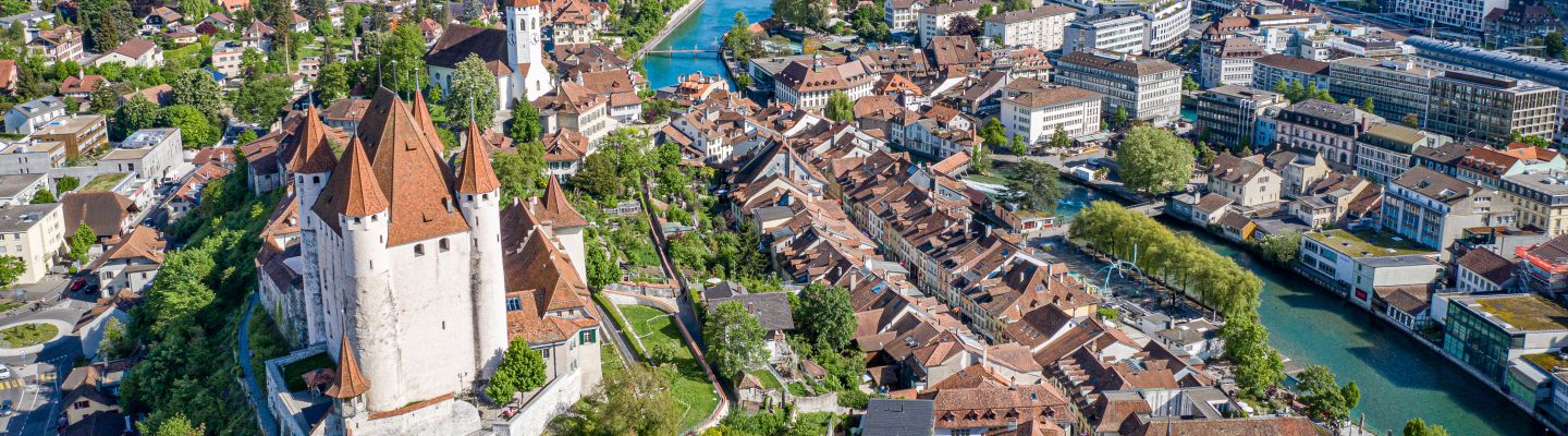 Beeindruckende Luftaufnahme von Thun, im Vordergrund das historische Schloss und die Schlosskirche. Der türkisfarbene Fluss schlängelt sich durch die Altstadt, im Hintergrund glitzert der Thunersee vor der imposanten Kulisse der Berner Alpen. Perfekte Kulisse für inspirierende Firmenevents in der Schweiz. ©Thun-Thunersee Tourismus Luftaufnahme von Thun mit Schloss und Alpenpanorama