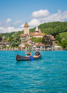 Zwei Personen paddeln entspannt in einem Kanu auf dem türkisblauen Thunersee. Im Hintergrund erhebt sich das historische Schloss Spiez mit seiner markanten Kirche, eingebettet in grüne Hügellandschaft. Ideale Szenerie für inspirierende Firmenanlässe oder Teamevents am See. ©spiez2017_by MikeKaufmann Kanufahrt vor Schloss Spiez
