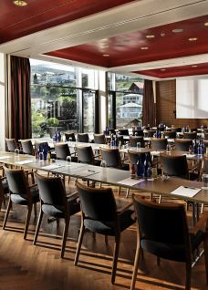 Heller Saal mit grossen Fenstern und einem einzigartigen Panoramablick auf die Spiezer Bucht. Warme Braun- und Orangetöne und hochwertiger Parkettboden sorgen für eine gemütliche Atmosphäre. ©Eden-Spiez Panorama