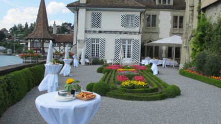 Auf der Schlossterrasse von Schloss Oberhofen stehen festlich mit weissen Tischdecken eingedeckte Stehtische, geschmückt mit Blumen und Apéro-Beilagen. Im Hintergrund erhebt sich das Schloss mit seinem berühmten kleinen Turm, der malerisch im Wasser steht. ©Schloss Oberhofen Schlossterrassen_hof_BrAn_389_