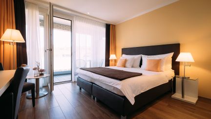 Modern eingerichtetes Doppelzimmer mit grossem Bett, stilvollen Leuchten und Zugang zu einem Balkon mit Glasfront. Helle Holzfussböden, dezente Farbgebung und viel Tageslicht schaffen eine einladende, entspannte Atmosphäre – ideal für Geschäftsreisende und Veranstaltungen am Thunersee. ©Hotel Schützen Komfortables Doppelzimmer mit Balkon