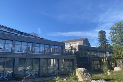 Panoramablick auf ein modernes Gebäude mit Glasfront, Aussenbestuhlung und Grünfläche – ideal für Meetings, Seminare und Events in der Region Thunersee. ©Seaside Spiez Moderne Seminar- und Tagungslocation mit Terrasse am Thunersee