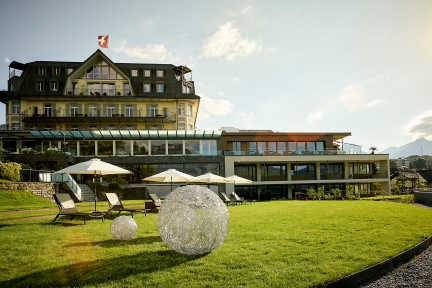 Blick auf ein Seehotel am Thunersee mit moderner Terrasse, Sonnenschirmen und gepflegter Rasenfläche – stilvolle Location für Meetings und Events. ©Strandhotel Belvédère Seehotel mit Terrasse und Eventrasen am Thunersee bei Abendsonne
