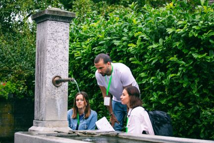 Drei Teilnehmende besprechen Unterlagen am Steinbrunnen im Grünen – informeller Austausch und Teamarbeit in der Region Thun-Thunersee. @FoxTrail Gruppe am Brunnen bei Outdoor-Event in Thun-Thunersee, mit Notizen und Namensbändern