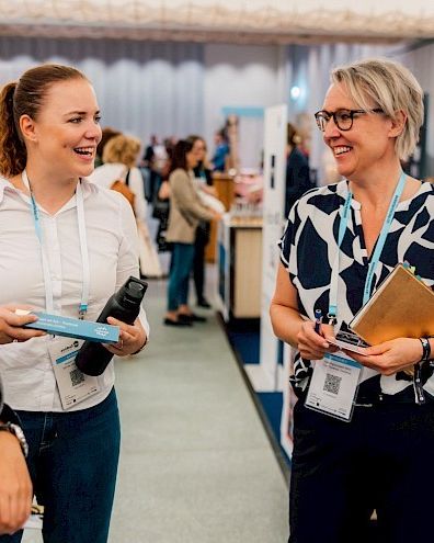 Drei Berufsfrauen unterhalten sich angeregt in einer modernen Kongressumgebung. Im Hintergrund sind weitere Teilnehmer und Messestände zu sehen. Diese Szene spiegelt die offene und inspirierende Atmosphäre für Geschäftstreffen und Events in der Region Thunersee wider. ©Assistants Day Zürich Persönliche Beratung von Thun-Thunersee Tourismus