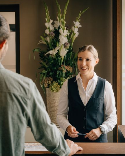 Eine freundliche Rezeptionsmitarbeiterin begrüßt einen Gast mit einem Lächeln an der Hotelrezeption. Im Hintergrund steht eine stilvolle Blumenvase, die dem modernen Empfangsbereich in Thun eine einladende Atmosphäre verleiht. Perfekter Start für einen Aufenthalt am Thunersee. ©Deltapark Herzlicher Empfang an der Rezeption