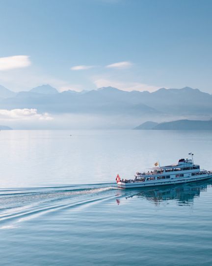 Ein Ausflugsschiff gleitet gemächlich über den ruhigen Thunersee, während sich im Hintergrund majestätische Alpen erheben. Die sanfte Morgensonne sorgt für eine beruhigende Atmosphäre und lässt das Wasser und die umliegende Landschaft in zarten Blautönen leuchten. ©BLS Schifffahrt. Schifffahrt auf dem Thunersee